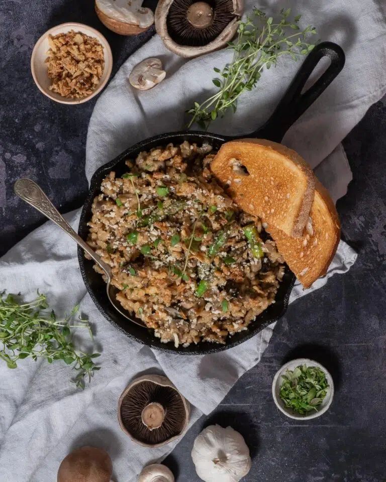 A top down view of a cast iron pan filled with creamy vegan mushroom risotto, sprinkled with fresh herbs and surrounded by herbs, fresh mushrooms and crispy fried onions