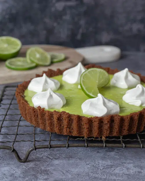 A vegan key lime pie with ginger nut base and tangy green lime filling sat on a metal rack, topped with piped whipped cream and a lime slice