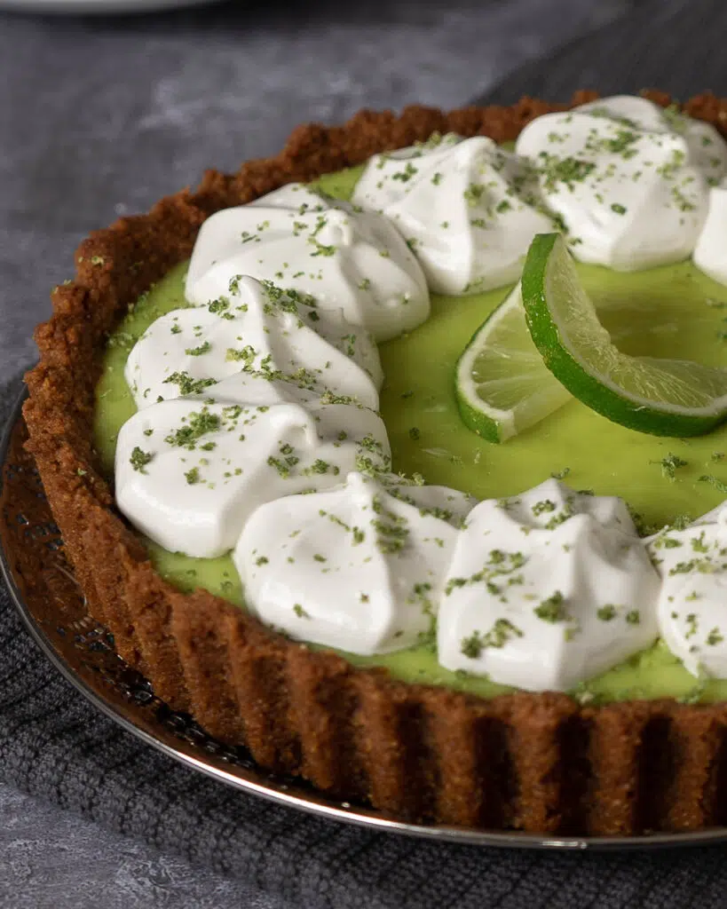 A vegan key lime pie with ginger nut base and tangy green lime filling, topped with piped whipped cream and a lime slice