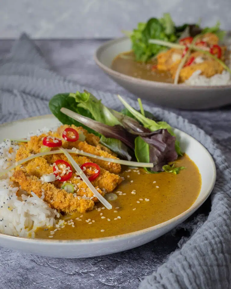 A bowl of vegan katsu curry with white rice and a meat free chicken fillet, with a garnish of salad, red chilli, spring onion and sesame seeds.