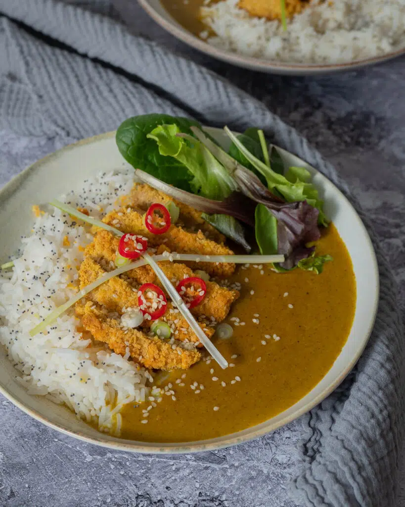 A bowl of vegan katsu curry with white rice and a meat free chicken fillet, with a garnish of salad, red chilli, spring onion and sesame seeds.