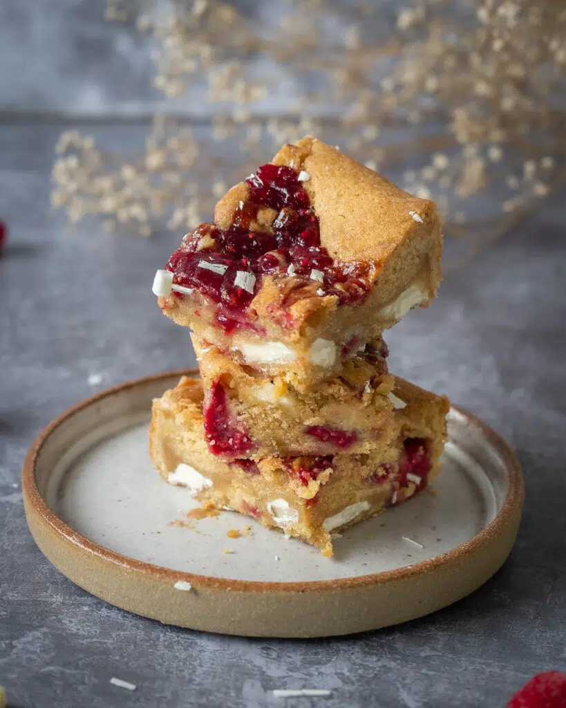 Three blondies stacked on a plate, showing the white chocolate, fresh raspberries and raspberry jam stirred through.