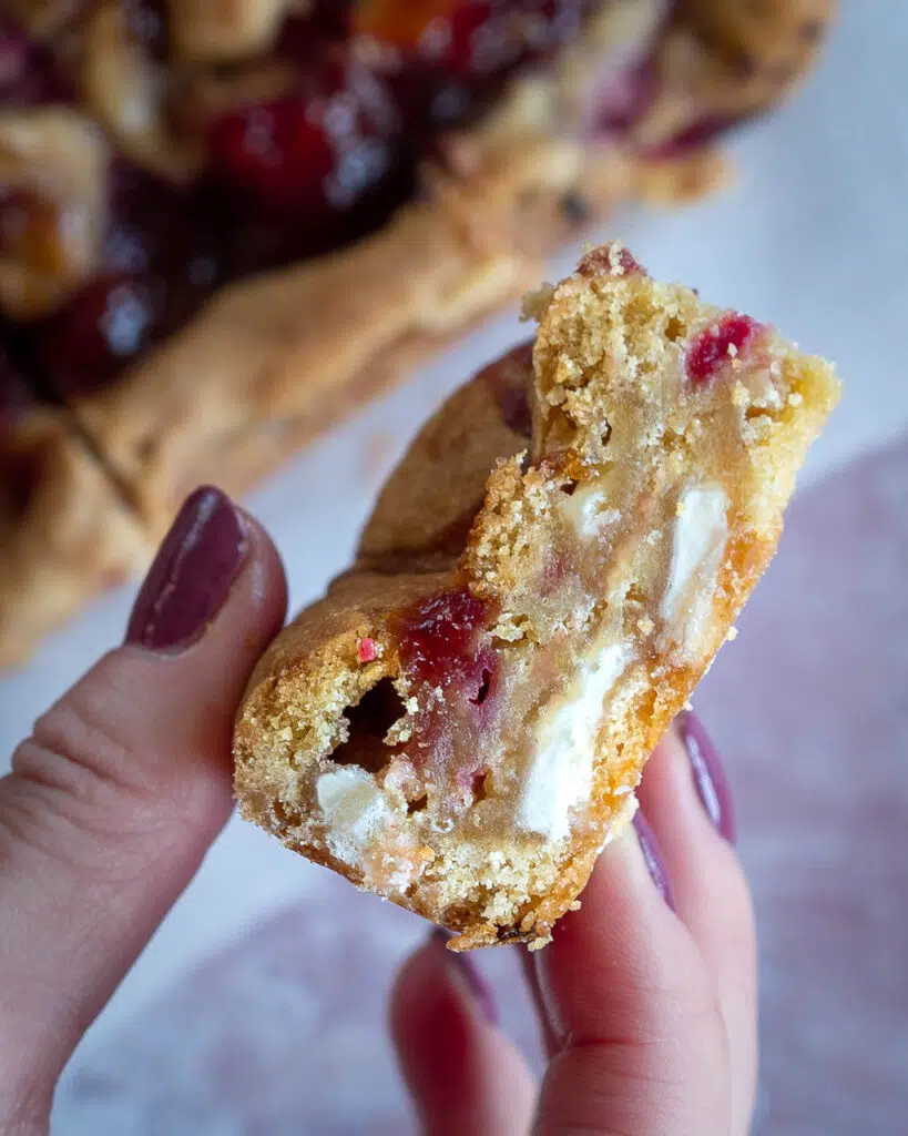 A hand holding a vegan blondie, displaying the chunks of white chocolate and fresh raspberries that have been baked into the centre.