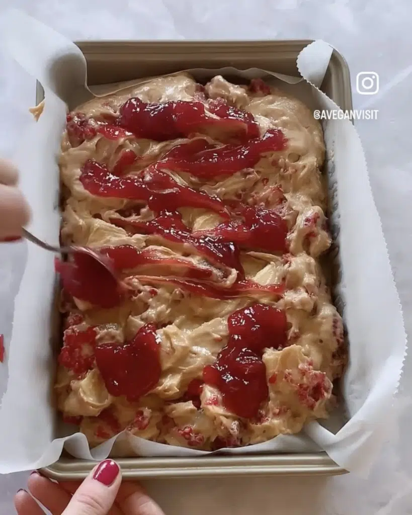 Raspberry jam being swirled on top of a brownie pan filled with blondie batter