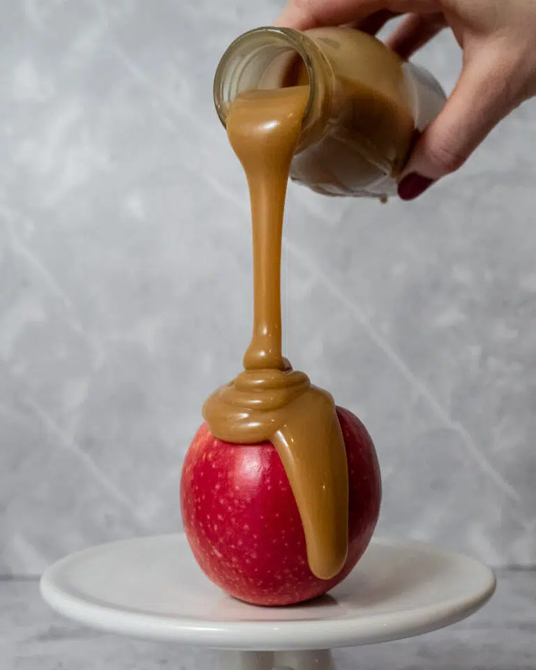 A red apple having creamy vegan caramel sauce poured over it, the thick sauce spilling down the sides.
