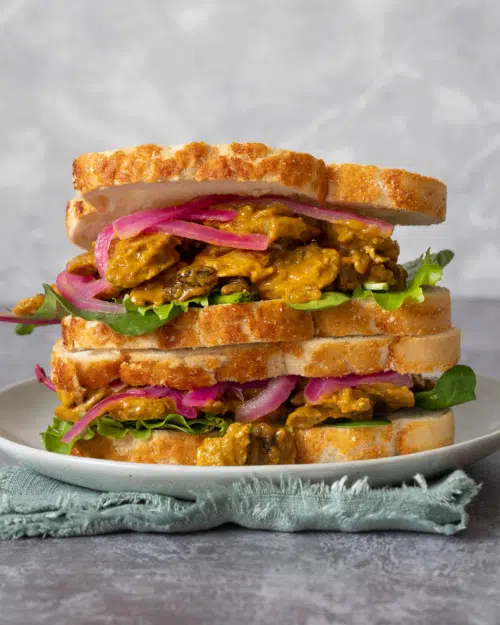 A stack of colourful vegan coronation chicken sandwiches with crusty farmhouse bread, coronation chick'n, lettuce and pickled red onions