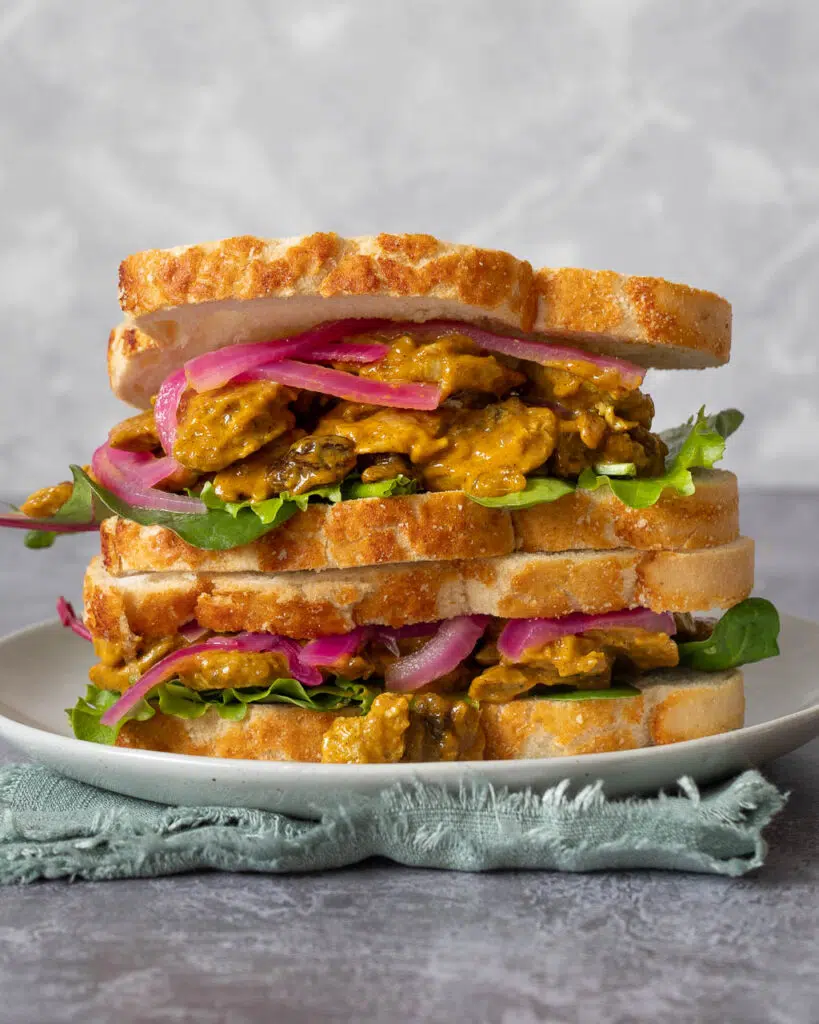 A stack of colourful vegan coronation chicken sandwiches with crusty farmhouse bread, coronation chick'n, lettuce and pickled red onions