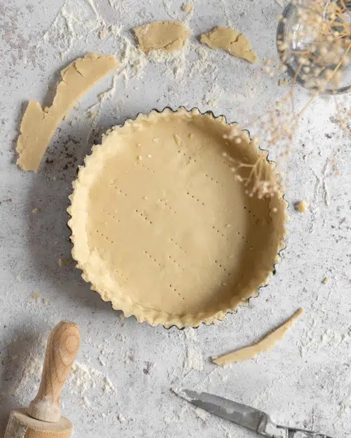 Sweet vegan shortcrust pastry lining a pie case, with offcuts surrounding it along with a knife and wooden rolling pin