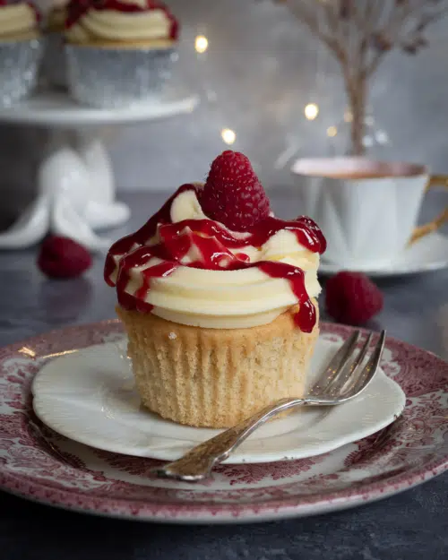 A pretty raspberry trifle cupcake sat on a stack of plates, with vanilla buttercream, a zigzag of raspberry jam and a fresh raspberry.