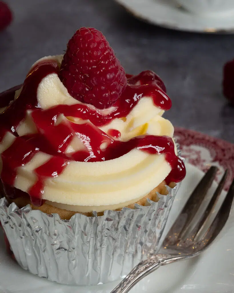 Close up of a pretty vegan raspberry trifle cupcakes sat on a stack of plates, with vanilla buttercream, a zigzag of raspberry jam and a fresh raspberry.