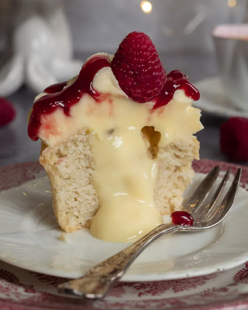 A vegan raspberry trifle cupcake sat on a white plate, cut in half with the custard centre spilling out