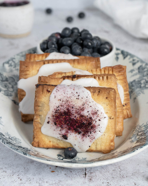 A oval white and blue floral dish holding a batch of freshly cooked blueberry vanilla vegan pop tarts, topped with icing and freeze dried blueberries.