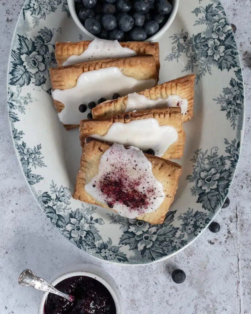 A oval white and blue floral dish holding a batch of freshly cooked blueberry vanilla vegan pop tarts, topped with icing and freeze dried blueberries.