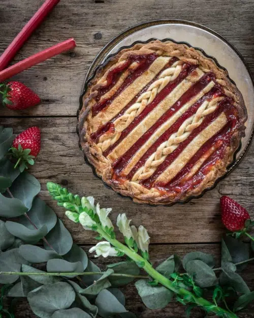 A flat lay of a pretty plaited strawberry and rhubarb pie surrounded by fresh strawberries, rhubarb sticks and green and white flowers and foliage