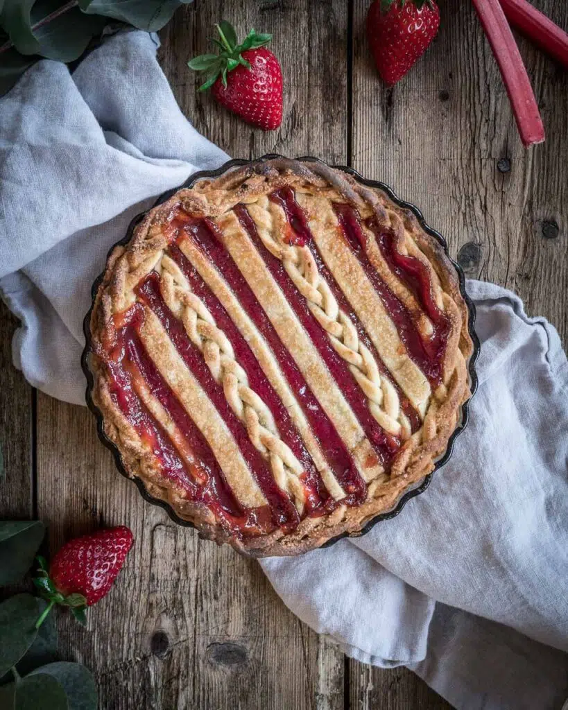 A top down photo of a round strawberry and rhubarb pie with lattice top, sat on a wooden table top with a grey napkin and fresh strawberries to decorate