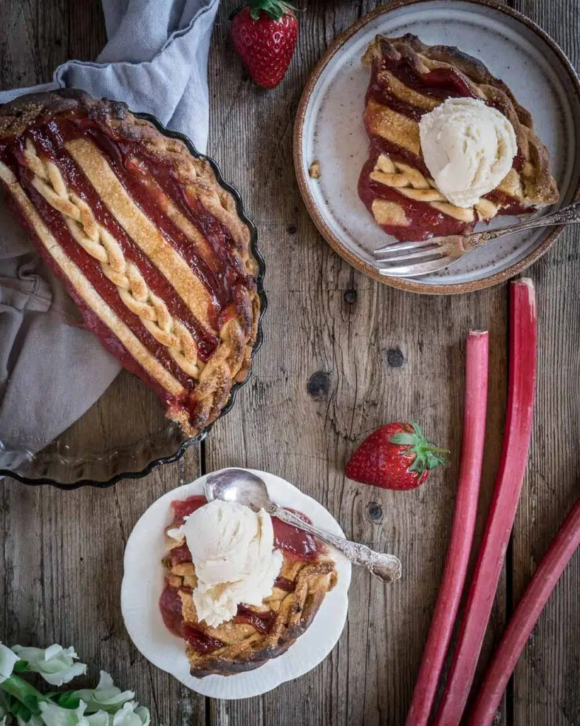 A wooden table top holding a rhubarb and strawberry pie with two plates dished up with vegan ice cream on top and fresh strawberries and rhubarb sticks on the table