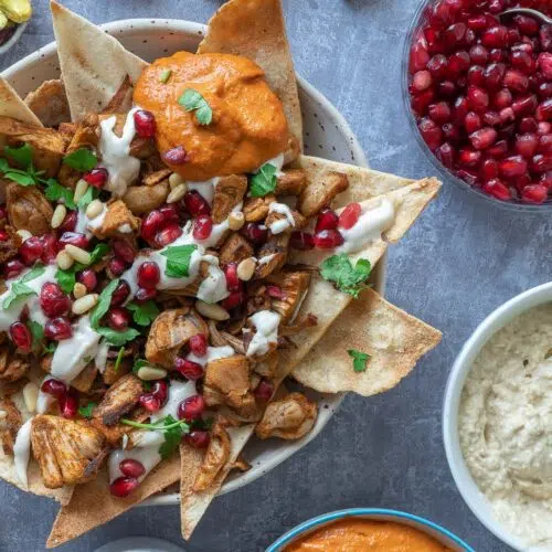 A close up of a colourful bowl of jackfruit fatteh loaded with pitta nachos, spiced jackfruit, pomegranate, tahini sauce, pine nuts, muhammara dip and fresh parsley.