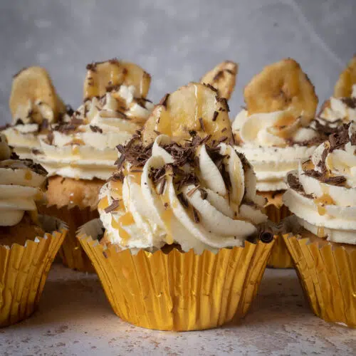 Three rows of vegan banoffee pie cupcakes in gold cupcake cases with whipped buttercream on top, a drizzle of caramel sauce, sprinkled dark chocolate and a banana chip