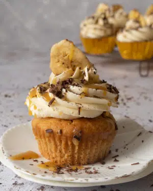 A vegan banoffee pie cupcake on a small white plate with whipped buttercream on top, a drizzle of caramel sauce, sprinkled dark chocolate and a banana chip
