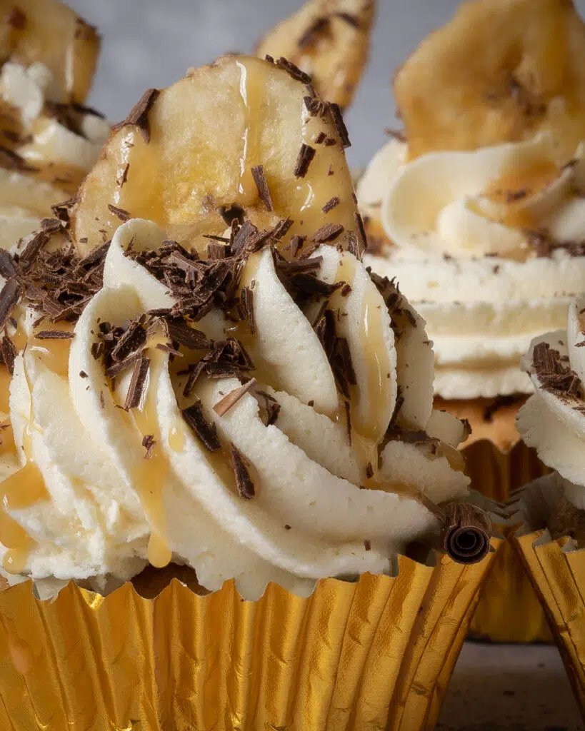Close up of vegan banoffee pie cupcakes in gold cupcake case with whipped buttercream on top, a drizzle of caramel sauce, sprinkled dark chocolate and a banana chip