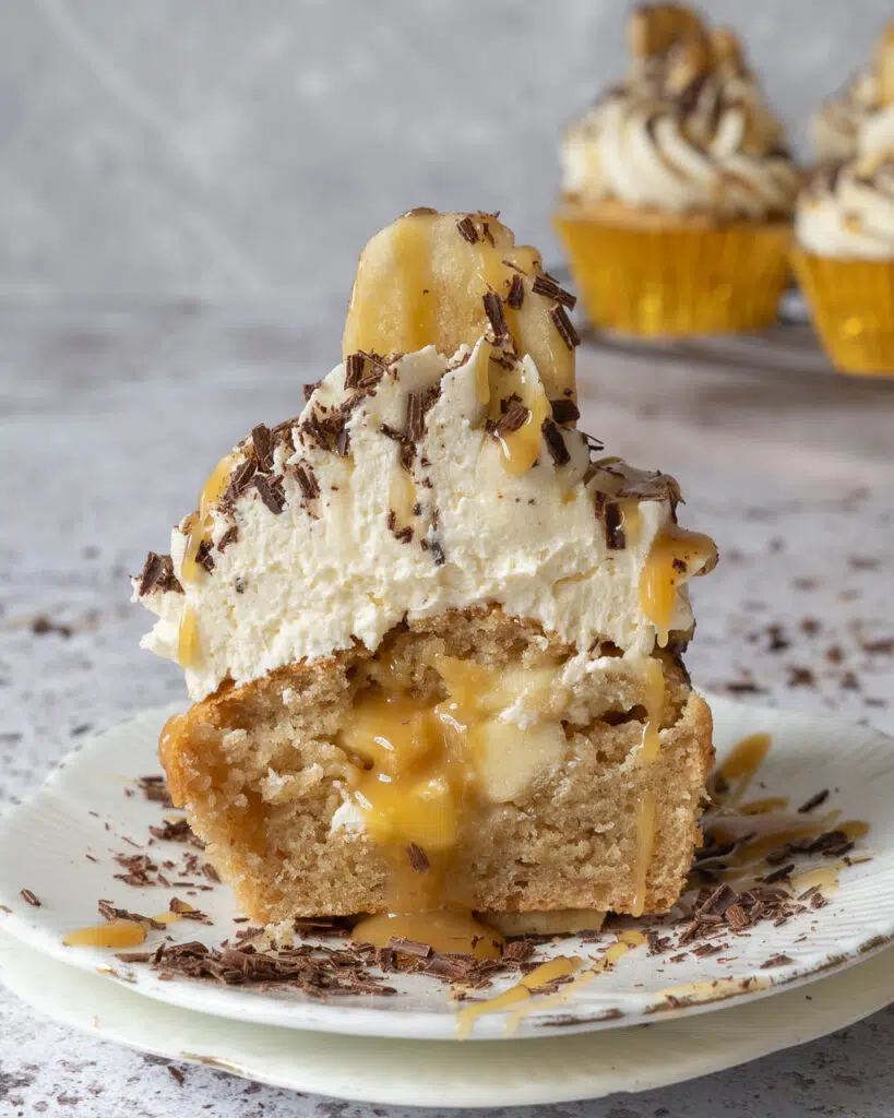 A vegan banoffee pie cupcake cut in half on a small white plate revealing a centre filled with diced banana and caramel sauce. The cupcake is topped with whipped buttercream, a drizzle of caramel sauce, sprinkled dark chocolate and a banana chip.