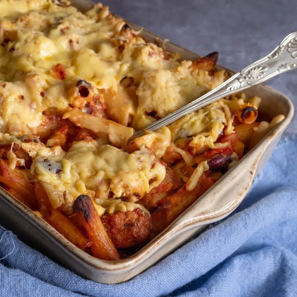 A baking dish filled with cheesy vegan sausage and bean pasta bake, with a serving spoon poised to scoop out a portion