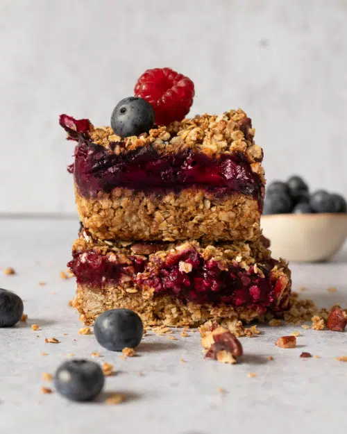 Two vegan berry crumble bars stacked on top of each other, revealing the layers of flapjack, jam and crumble, with fresh berries on top