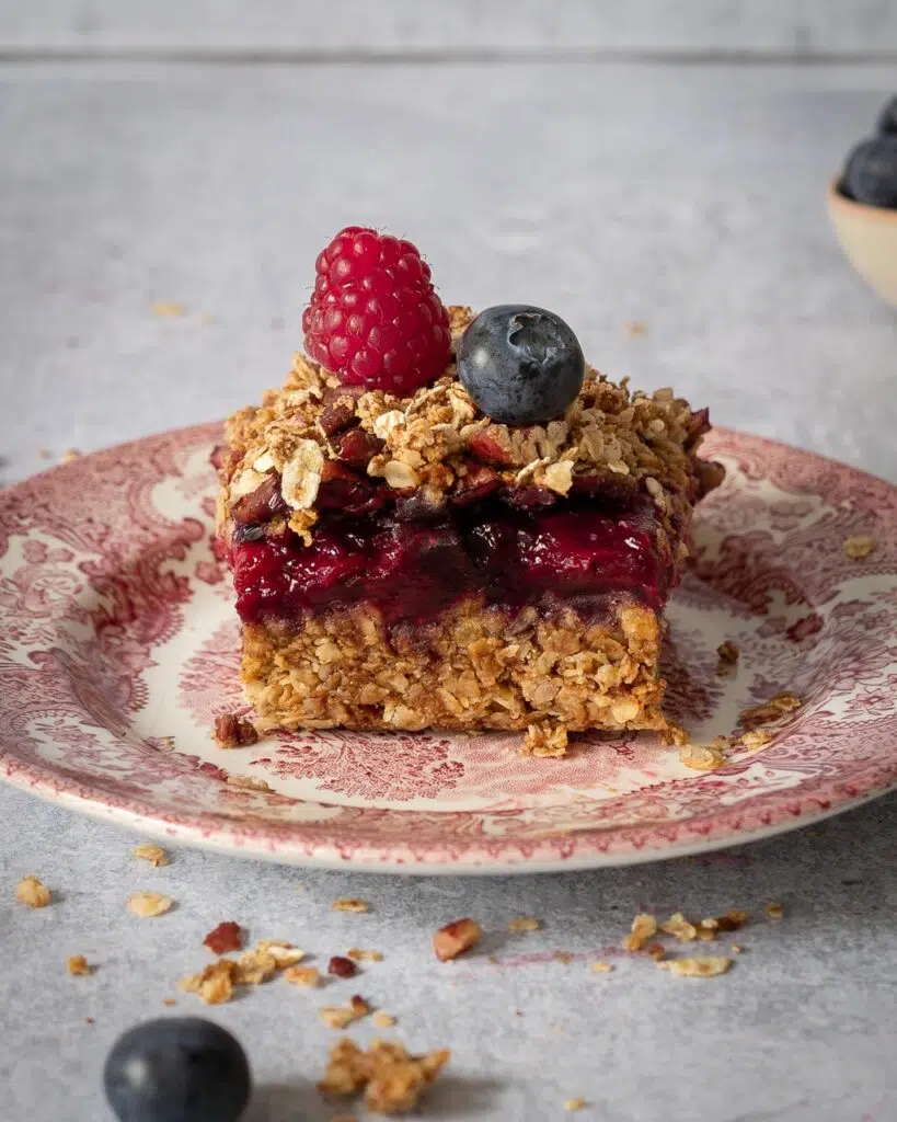 A vegan summer berry crumble bar on a pretty pink and white plate ready to be eaten, showing the layers of flapjack, berry jam and pecan crumble, with fresh berries on top