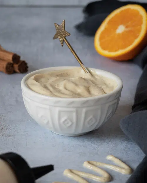 A bowl of creamy tofu icing being stirred by a gold star spoon, with an orange cut in half resting beside it along with cinnamon sticks