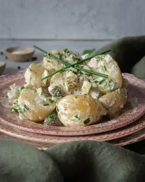 A stack of pretty pink and white plates piled with vegan potato salad in a creamy sauce, topped with fresh chives