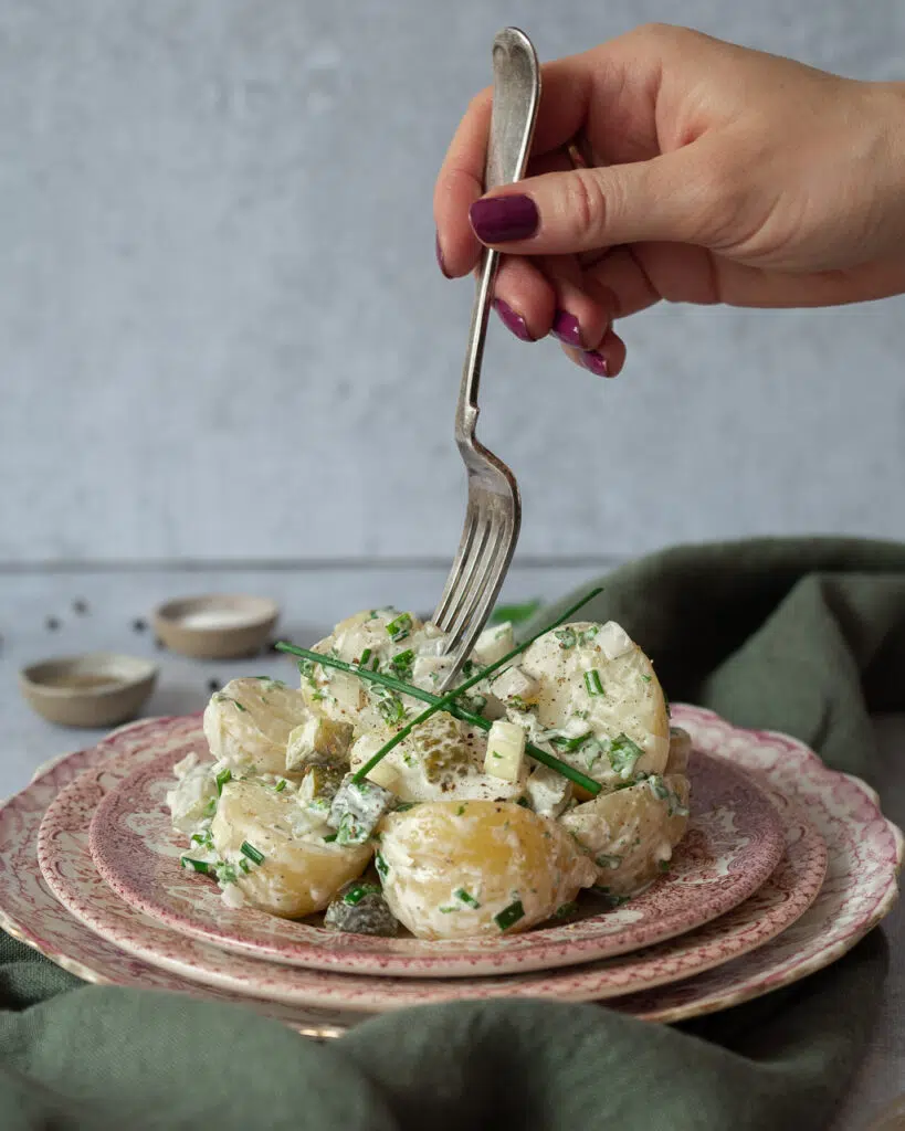 A person sticking a fork into a pile of freshly made vegan potato salad, in a creamy sauce with fresh herbs sprinkled on top