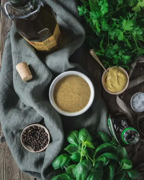 A flat lay of a bowl of healthy vegan salad dressing surrounded by all of the ingredients used to make it including fresh herbs, peppercorns, olive oil and maple syrup