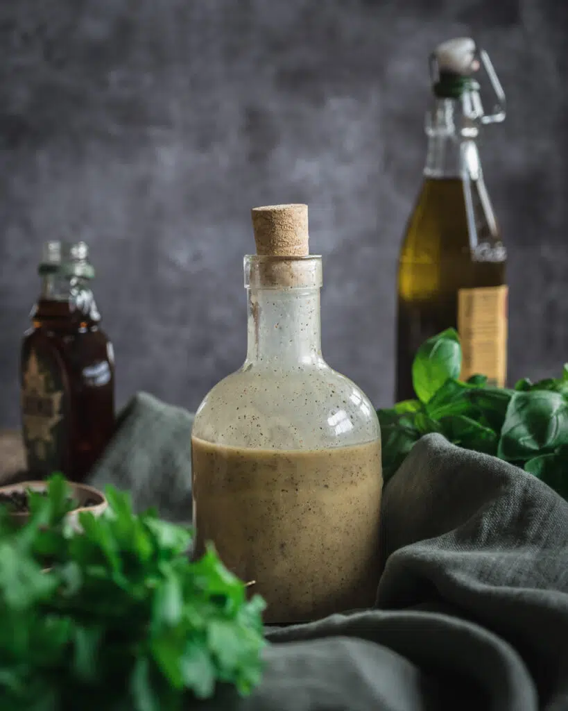 A glass bottle with cork stopper filled with healthy vegan salad dressing, with an out of focus bottle of olive oil and maple syrup behind and fresh herbs in the foreground