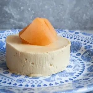 A creamy yellow round cheesecake on a blue patterned plate with a folded piece of orange melon on top.