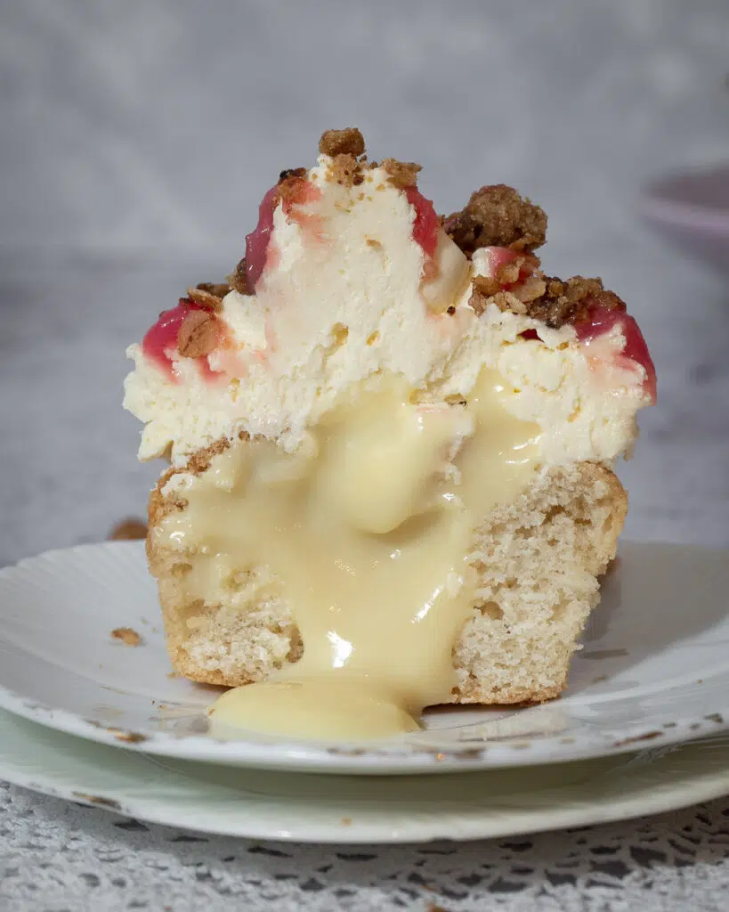 A vegan rhubarb crumble cupcake on a small white plate, cut in half to reveal a dairy free custard centre oozing out