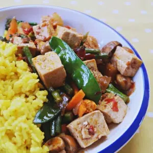 A bowl of vegan stir fry with cubes of firm tofu, green fresh mange tout, carrot strips and chilli flakes next to bright yellow saffron rice.
