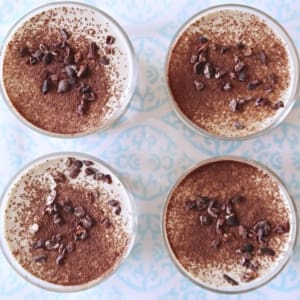 Homemade raw vegan tiramisu. Birds eye shot with four round dishes sprinkled with cocoa and cacao nibs on a blue and white table cloth.