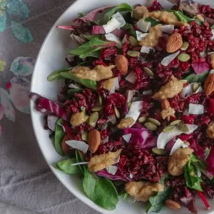 A round bowl of salad with a bed of leaves, red cabbage, almonds, dressing and cranberries on a floral table cloth.