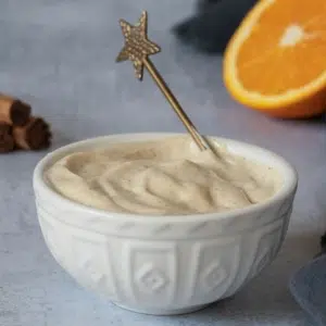 Silky smooth orange and cinnamon tofu icing in a white bowl with a gold star spoon. With fresh orange and cinnamon sticks out of focus behind.