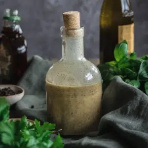 A glass bottle of salad dressing with a cork stopper. Surrounded by an olive green linen napkin, a bottle of maple syrup and olive oil and fresh herbs.