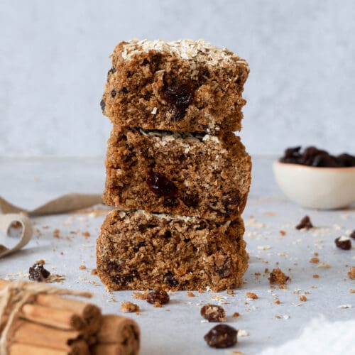 A stack of cinnamon raisin elevenses cake bars with a raisins scattered around and a pile of cinnamon sticks out of focus in front