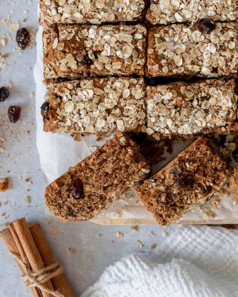 Top down view of a batch of cinnamon raisin elevenses cake bars on a chopping board, having been cut into slices, sprinkled with oats on top and raisins scattered around