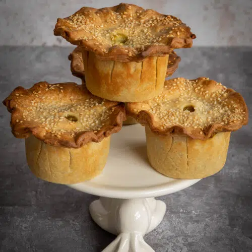 Four pretty curried chickpea and potato vegan pies piled on a cake stand, sprinkled with sesame seeds