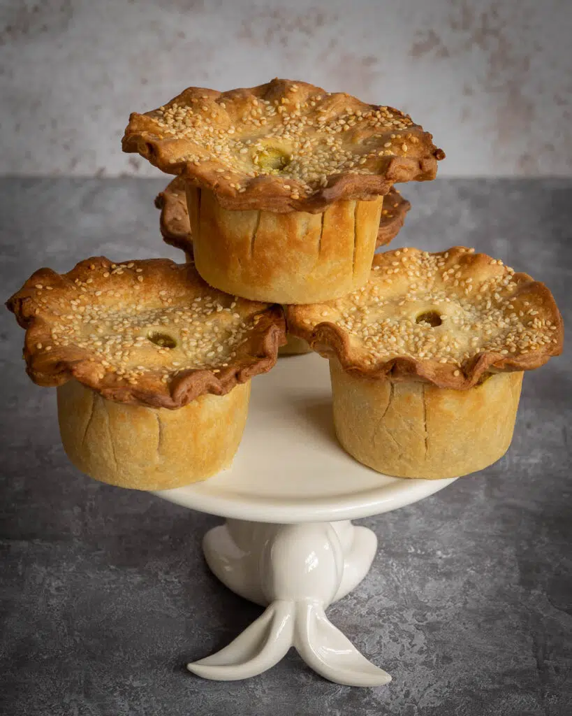 Four pretty curried chickpea and potato vegan pies piled on a cake stand, sprinkled with sesame seeds
