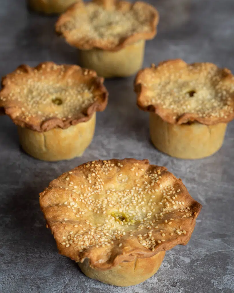 Four curried chickpea and potato hot water crust vegan pies lined up, with crimped lids and sprinkled with sesame seeds