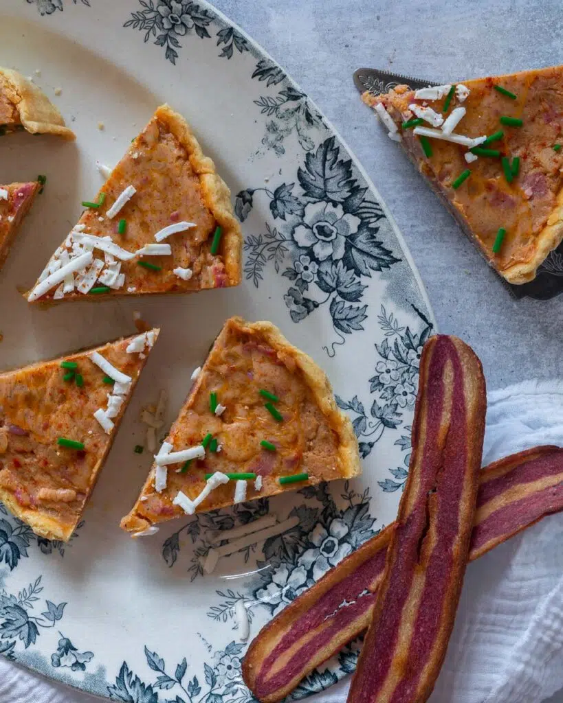 A birds eye view of an oval serving dish with slices of vegan Quiche Lorraine on it, sprinkled with grated cheese, fresh chives and vegan bacon