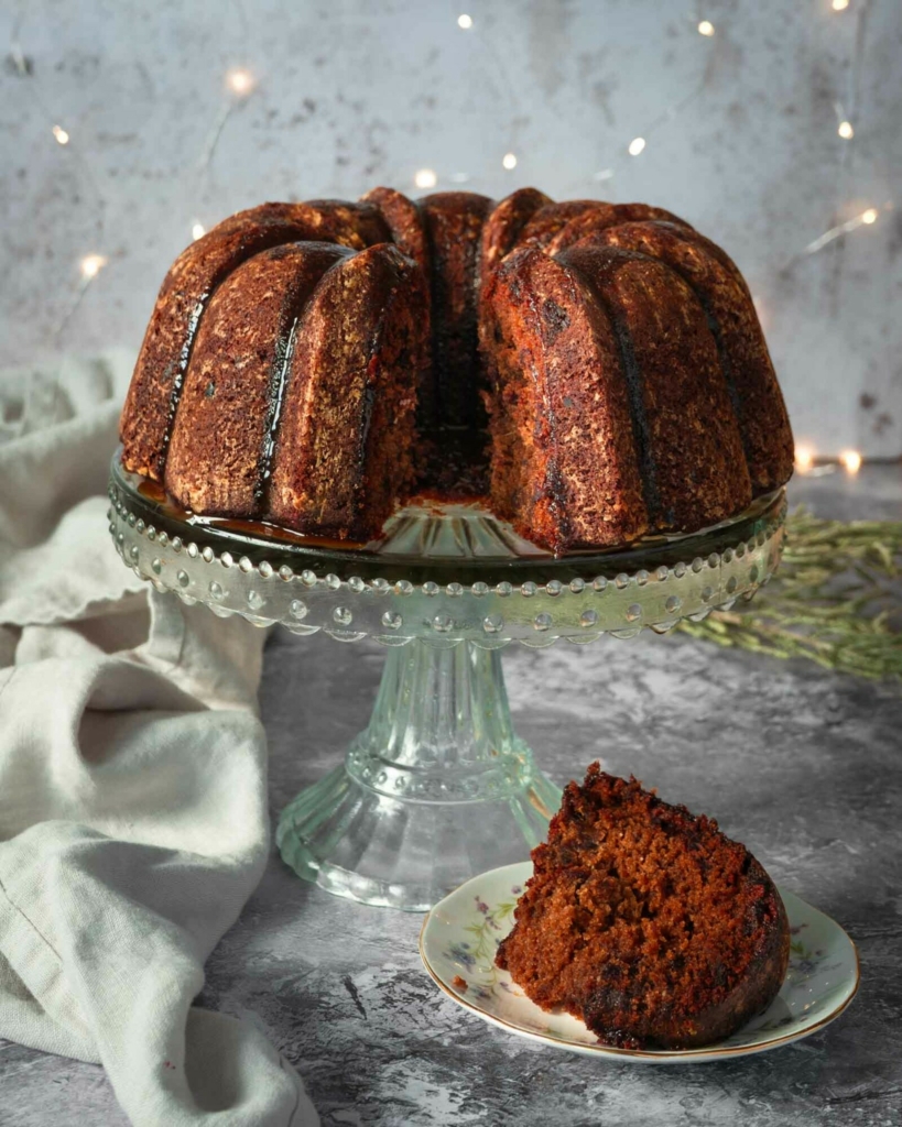 A richly coloured sticky toffee pudding cake in the shape of a ring, sat atop a glass cake stand, with toffee sauce running down the sides.