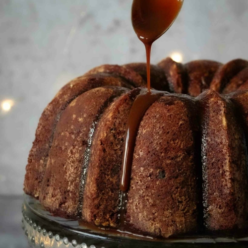 A vegan sticky toffee pudding cake on a glass cake stand with toffee sauce being drizzled from a spoon on top