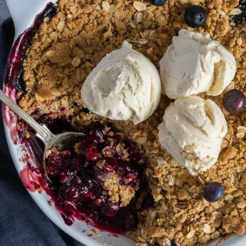 A large round dish of blueberry and thyme crumble with three scoops of vegan vanilla ice cream on top