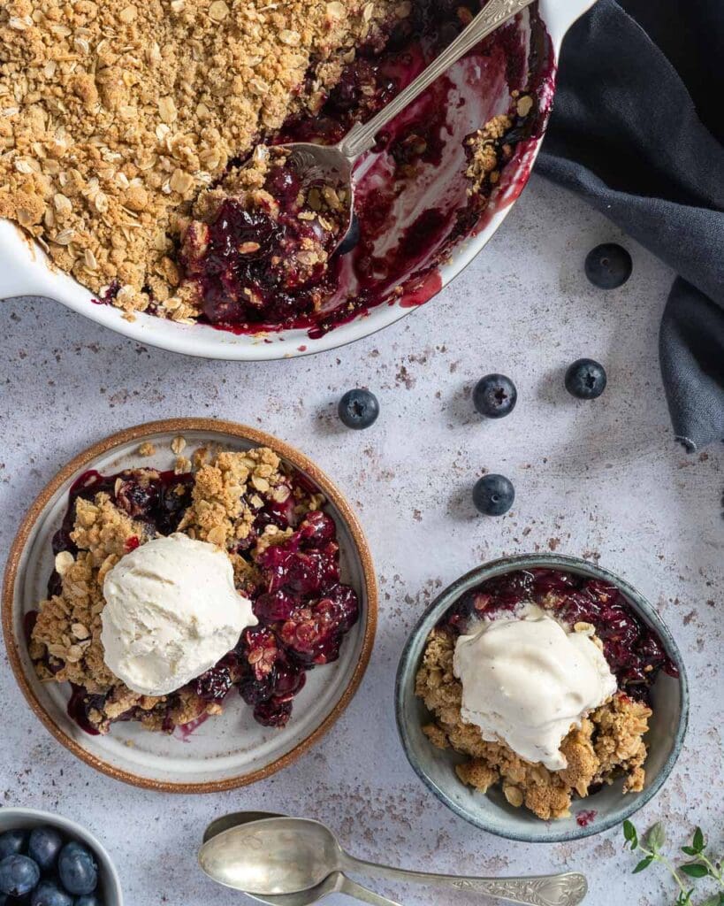 Two plates of rich purple blueberry thyme crumble with a scoop of vegan vanilla ice cream on top